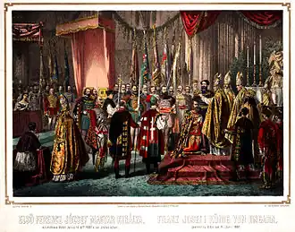 A bishop puts a crown on the head of a bearded man kneeling before him in a crowded large hall