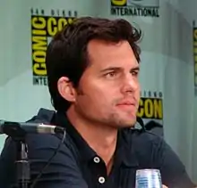 Color photograph of a white man with brown hair sitting in front of a ComicCon poster.