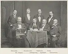 Black and white photo; seven men in suits sit and stand around a table