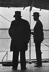 Kruger viewed in silhouette from behind, Bredell to his right. Kruger is wearing his top hat.
