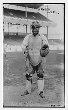 A man wearing a baseball uniform and full catcher's gear (facemask, catcher's mitt, chest protector, and leg guards) stands poised–catch a ball.