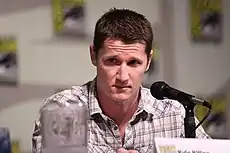 A man with short brown hair and a buttoned shirt in front of a microphone.