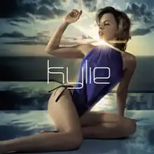 An image of Minogue posing with the blue sky and sea in the background, wearing a blue swimsuit and raising her right hand