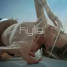 An image of a woman lying down on a marble-like surface, wearing a tan-coloured bikini. The name "Kylie" is superimposed on her.