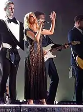 Minogue singing on stage in a brown dress surrounded by male dancers in tuxedos