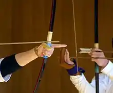 a hand holds a bow while the complementary hand, wearing a glove, draws the bowstring (an arrow is nocked); in the background another pair of hands gestures to places on another bow
