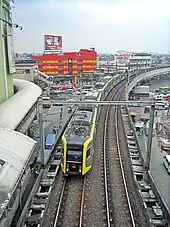 Below, on elevated tracks surrounded by a daytime urban landscape of roads, cars, buildings, and billboards, a metro train approaches, its front driver's window framed on the sides by angled planes and yellow markings visible as the rest of the train trails behind curving slightly to the right into the distance.