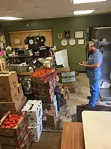 many stacked boxes of tomatoes and other produce in a cramped kitchen, and a man clad in overalls writing on a clipboard