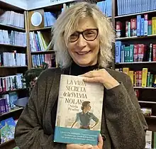 Color photo of a woman holding a book, the bookshelves behind her filled with other books.