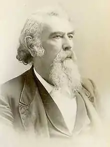 A man with white hair and a white beard and mustache, facing right. He is wearing a white shirt, black vest, and black jacket