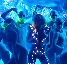 A blond woman dancing in a blue dress, which has small glowing squirrels on it. She is surrounded by dancers in silver, body-hugging dress with a neon green mustard in front