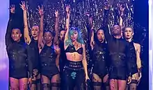 A group of people standing with their right hand raise up. They are decked in black garments with bare legs. Central to them is a woman who has blue hair and tufts of hair from her underarms and at her pubic area.