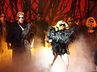 A group of people stand on a stage in black, feathery dresses and black sunglasses. Prominent among them is a blond woman, with a mouthpiece attached to her ear. Behind the group, a red background can be seen, interspersed with black thorn-like structures.