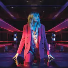 Ladyhawke in a pink suit looking to her left while stepping into an indoor pool in a somewhat colourful but darkened room