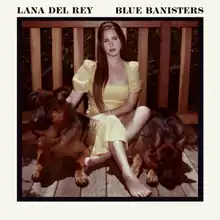 A picture of woman in a yellow dress sitting with two dogs by her sides on a wooden porch in front of the banisters. The picture is surrounded by a white outline with the text "Lana Del Rey" and "Blue Banisters" written in black.