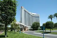 Distant ground-level view of a curved building with a white, concrete facade and dark, blue-tinted windows. Monorail tracks are visible in the foreground.