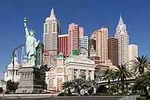 Ground level view of a 50-story building that looks like a city skyline, forming several different towers of varying heights. A prominent copper statue holding a torch is visible at the front of the image. A roller coaster is also visible between the statue and the towers.