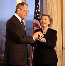 Hillary Clinton standing with Russian Foreign Minister Sergey Lavrov. Both of them are holding a "reset button". They are in a room with a window to the left and an American flag behind them