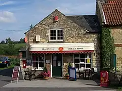 Stonebuilt shop with modern PVC folding canopy, on a sunny day. A bright pot of flowers on the left, and ivy up the stone building on the right.