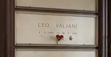 A white marble gravestone on the wall of a chapel, with only the name and dates of birth and death inscribed, a small photograph of the deceased and a bunch of red carnations