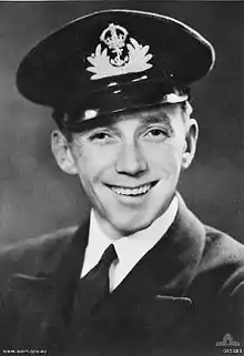 A head-and-shoulders portrait of a smiling man in naval uniform. He is wearing a cap that has a badge on the front depicting a crown that has an anchor hanging off it in the middle of splashing waves.