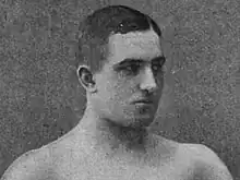 Monochrome portrait of Leslie Hood taken in the studios of Gale and Polden in 1901. Hood is photographed with short hair, bare shoulders, and looking to his left. The photograph was used to illustrate the effectiveness of Eugen Sandow's course on building muscular strength.