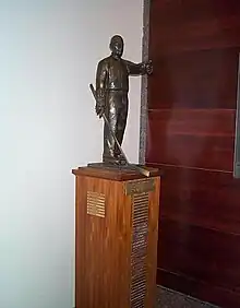 Bronze statuette of a man coaching hockey while wearing skates and holdindg a stick, mounted upon a tall wooden plinth with name plates for each recipient of the trophy.