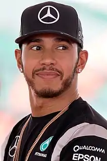 Lewis Hamilton looking to the left to the camera is wearing a black baseball cap, a gold chain necklace and a black T-shirt with sponsors logos