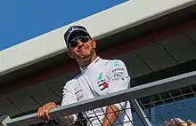 Lewis Hamilton wearing a black baseball cap and silver racing suit looking to the crowd