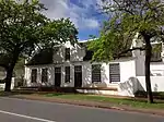 A short distance from the railway station at the corner which Dorp Street forms with Somerset West Road, stands the first remarkable old house in this street. It is called Libertas Parva or Little Libertas and it is on the right side of the street (now Rembrandt van Rijn Art Centre). The oldest feature of the house is its four side gables; they are of the holbol type, but show a breaking up of the concaves and convexes into wavy lines reminiscent of a similar tendency in the front gables of the Zevenrivieren-Hazendal type, c, 1790. The house may therefore date from 1783 or soon after. Philip Jacob Haupt purchased the farm in 1819 and it must be accepted that he was responsible for the erection of the present front gable, with almost straight outlines, with the two front doors built in and also for the Georgian windows. Their outer surrounding pilasters are continued upward to form the outer gable pilasters. The Libertas Parva building complex is a fine example of the Stellenbosch architecture in the 18th and 19th centuries and lends a particular character to that part of the village. As the home of well-known Stellenbosch families it is also of great historic value.