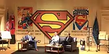 Three men seated onstage, flanked by Superman material