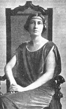 A white woman, seated, wearing a headband across her forehead and a loose-fitting velvety tunic-style dress