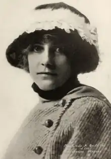 A white woman wearing a hat with a brim and feather embellishment; she is also wearing a coat with an off-center buttoned closure