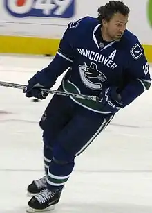 Hockey player in dark blue Vancouver uniform. He leans forward, holding his stick aloft.