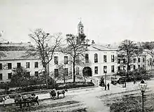 A black and white photograph, showing a stately-looking two storey building with white walls, extending out of shot to the left and right, with an arched cart entrance at the centre. A modest clocktower rises above the entrance, and the building is surrounded by neat shrubbery and iron railings. A wide street crosses left-right outside of the fence, with a handful of horse-drawn carts and pedestrians in 19th century clothing.