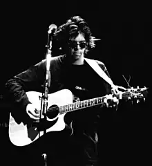 A black-and-white photograph of a man in black sunglasses performing on a stage with an acoustic guitar.