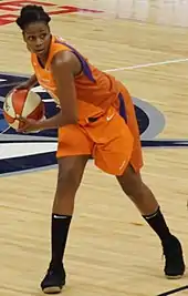 Young woman wearing orange basketball uniform leaning to her right carrying the ball