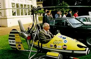 Small, one man, open-cockpit helicopter on a lawn about the size of a car next to it, with a man sitting in it.