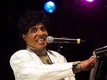 An African American man singing into a microphone, the microphone is being held up by a stand. The male is wearing a cream color suit that has black pin stripes.