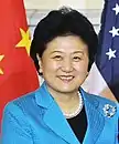 a smiling, aged woman, wearing a blue dress (and a black shirt under it), white necklace and standing in front of the American and Chinese national flags