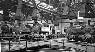 The inside of an engine shed with part of a turntable in the foreground. A number of engines are arranged round the turntable, each with their chimneys under large ventilation cowls.