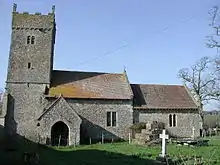 A square stone three step cross base, supporting a socket stone, in front of a small stone church and tower
