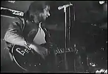 Upper body shot of a man playing a guitar with letters G-e-o visible. His head is turned partly to his left. There is a microphone stand in front of him. Other musical and studio equipment is around him. Another musician is obscured at bottom right.