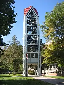 A three-dimensional vertical structure perhaps 70 feet (21 m) tall and 10 feet (3.0 m) wide by 10 feet (3.0 m) long is filled with tiers of large metal bells. The structure, standing in front of a large institutional building in a park-like setting, is supported by several metal legs. A sidewalk passes through the structure, which is open at the bottom to a height of perhaps 10 feet (3.0 m).
