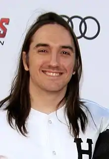 A white man is standing in front of a white, logo-covered wall; he has dark hair and is both facing and smiling into the camera.