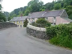 single-track tarmac road curving to the right over a stone bridge with a row of two-storey stone cottages beyond