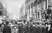 Black and white photograph of a crowded street, taken at eye-level. Clothes can be seen hanging at stalls along the sides of the street.