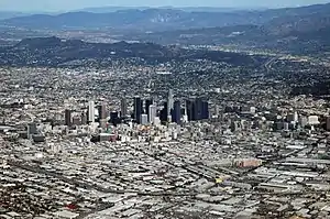 An aerial view of a widespread built-up area, skyscrapers in the central district, with mountains in the background.