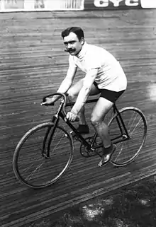 A black-and-white photograph of a man with three-colored sweater and shorts with a mustache sitting on a bicycle.