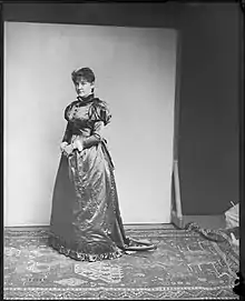 A woman with a serious expression stands in a high-collared, floor-length dress